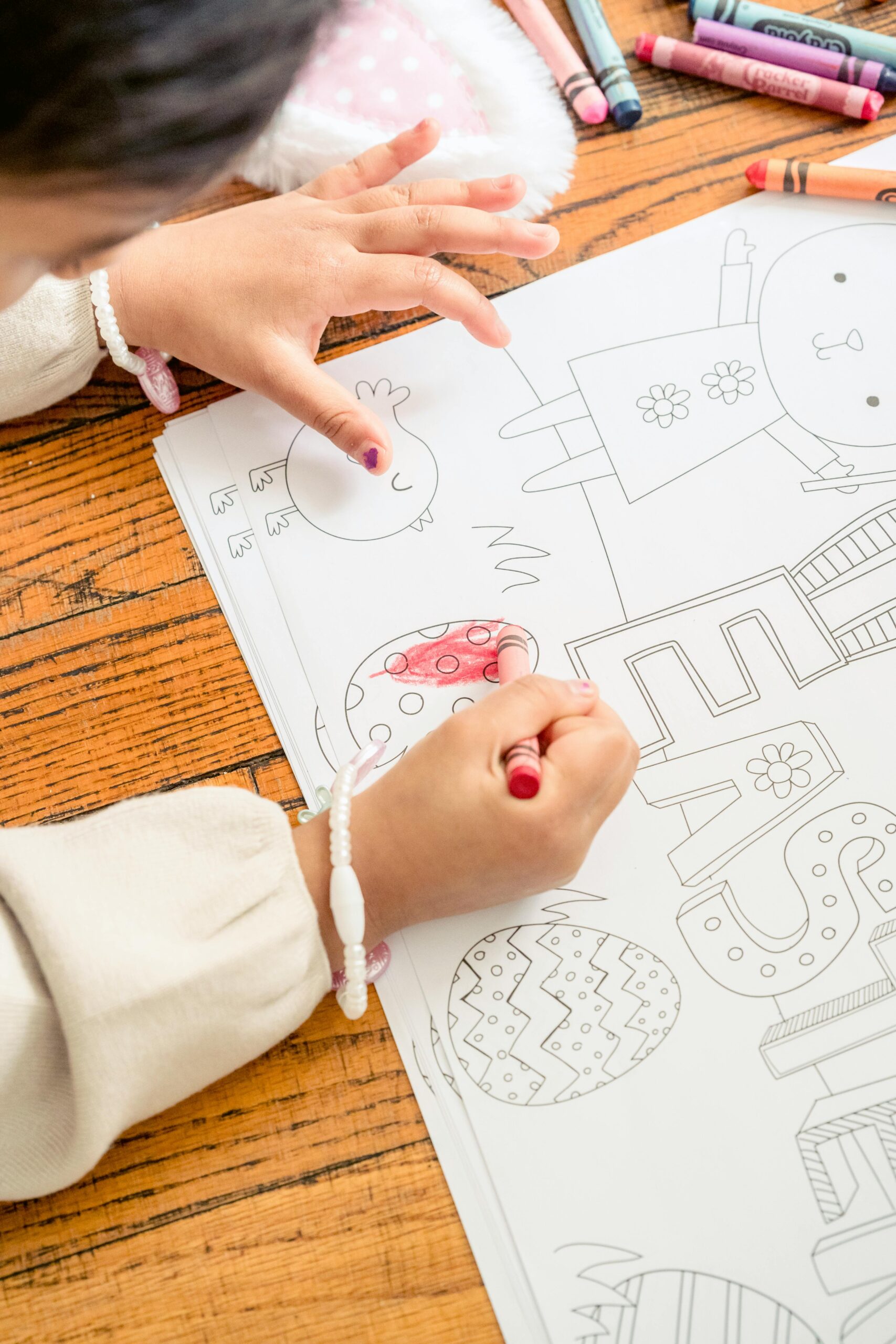From above of crop faceless kid in casual clothes sitting on wooden floor with wax pencil in hand and coloring paper with Easter word and eggs in light room